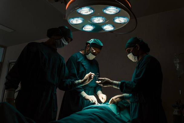 Zespół chirurgiczny wykonujący operację pacjentowi na sterylnej sali operacyjnej. W sali operacyjnej oświetlonej lampą, profesjonalny i pewny siebie zespół chirurgiczny zapewnia opiekę medyczną nieprzytomnemu pacjentowi. - Zdjęcie, obraz