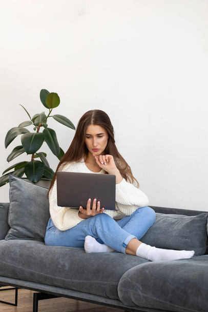 魅力的なブルネットの女性は自宅のソファに座ってインターネットで情報を勉強しています。女は膝の上に寝そべっているノートパソコンを見る。オンライントレーニング。リモートワーク. - 写真・画像