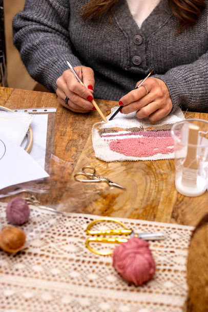 Embroidery punch needle workshop. Middle-aged woman with red painted nails applies glue on embroidered fabric. - 写真・画像