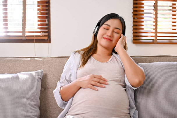 Relaxed and happy Asian pregnant woman in comfy clothes listening to music through her headphones while resting in her living room. - Foto, afbeelding