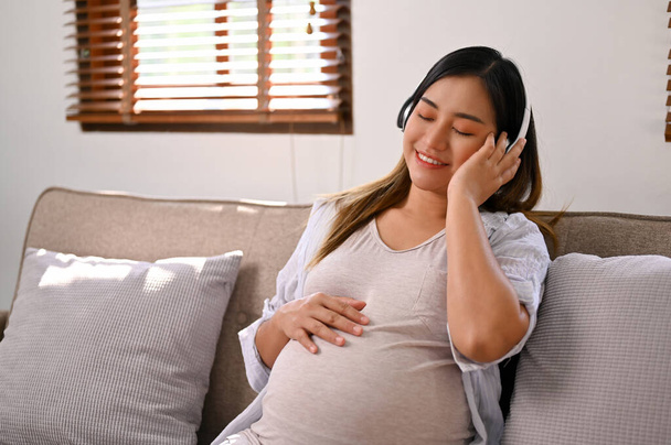 Happy Asian pregnant woman listening to music through her headphones on sofa in her living room. pregnancy lifestyle concept - Foto, Imagen
