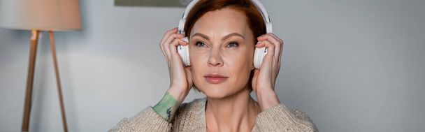 Redhead woman in cardigan using wireless headphones at home, banner  - Foto, imagen