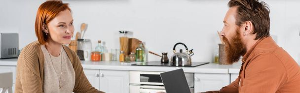 Улыбающаяся рыжая женщина смотрит на бородатого мужа рядом с ноутбуком на кухне, баннер  - Фото, изображение