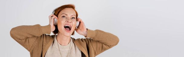 excited and red haired woman in headphones singing isolated on grey, banner  - 写真・画像