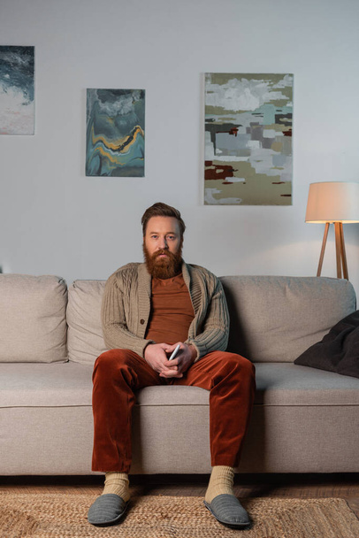 Evde kanepede otururken elinde akıllı telefon tutan hırkalı sakallı adam.  - Fotoğraf, Görsel