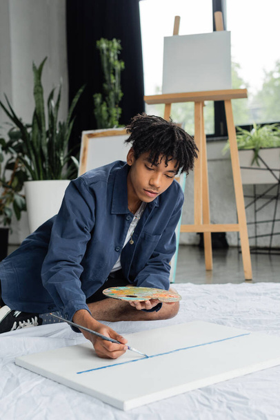 Young african american artist painting on canvas while sitting on cloth in studio  - 写真・画像