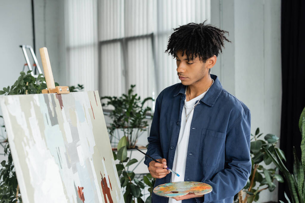 Junge afrikanisch-amerikanische Künstlerin mit Pinsel und Palette in der Nähe der Malerei im Atelier  - Foto, Bild