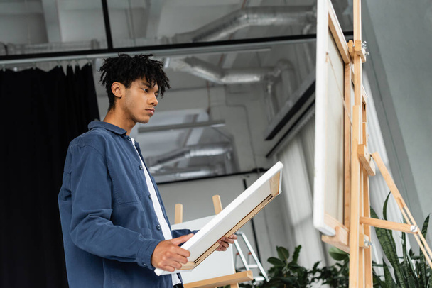 Low angle view of african american artist holding canvas near easel in studio  - Photo, Image