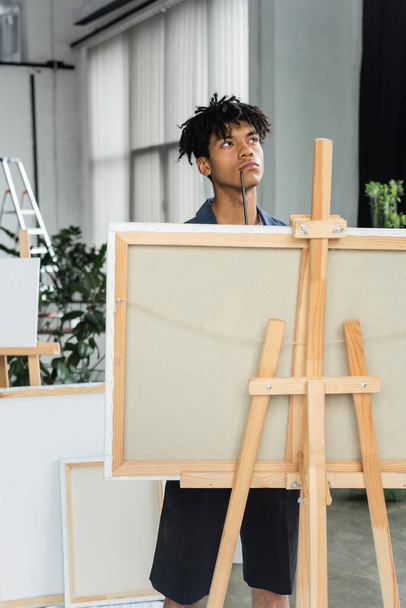 Pensive african american artist with paintbrush standing near canvas in studio - Foto, Bild