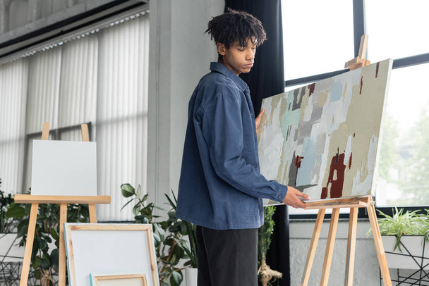 Young african american artist taking painting from easel in workshop  - Φωτογραφία, εικόνα