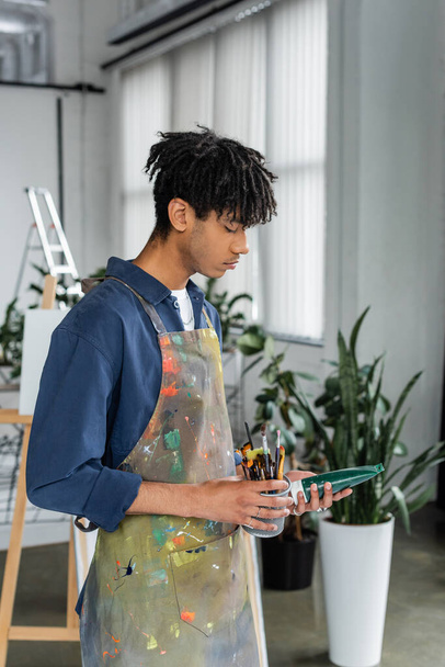 Vue latérale du jeune artiste afro-américain dans un tablier tenant des pinceaux et de la peinture en atelier  - Photo, image