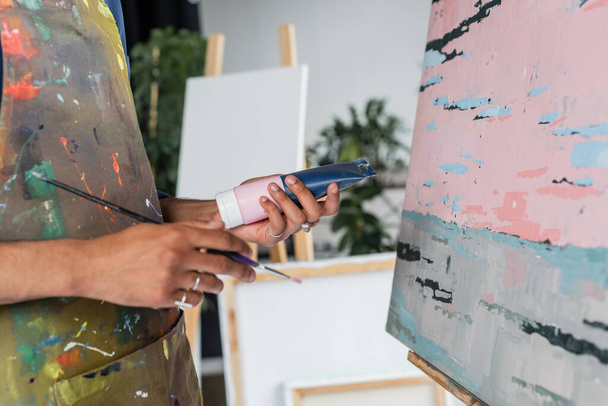 Обрезанный вид африканского американского художника в фартуке, держащего краску и кисть рядом с рисунком  - Фото, изображение