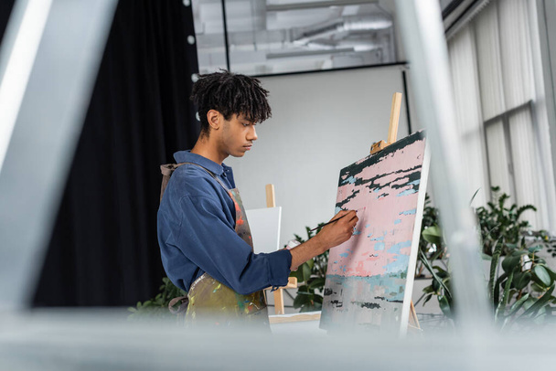 Vue latérale du jeune artiste afro-américain dans la peinture de tablier sur toile  - Photo, image