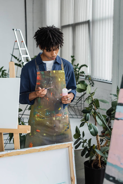 Artista afroamericano en delantal sosteniendo pincel y pintura cerca de lienzos en taller  - Foto, Imagen
