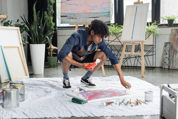 Artiste afro-américain tenant de la peinture près de la toile et pinceaux sur tissu en atelier  - Photo, image