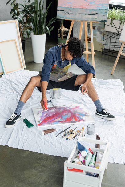 Νεαρός αφροαμερικανός καλλιτέχνης ρίχνει χρώμα σε καμβά κοντά σε χρώματα στο εργαστήριο  - Φωτογραφία, εικόνα