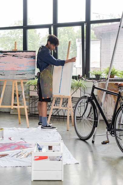 Seitenansicht eines jungen afrikanisch-amerikanischen Künstlers in Schürze, der Leinwand auf Staffelei in der Nähe von Fahrrad in der Werkstatt legt  - Foto, Bild
