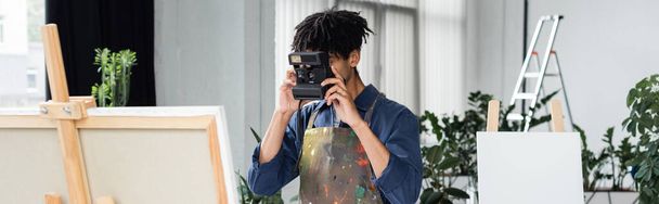 Artista afro-americano tirando foto na câmera vintage perto de tela no cavalete em estúdio, banner  - Foto, Imagem