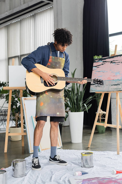 Artista afroamericano tocando guitarra acústica cerca de pinturas y caballetes en taller  - Foto, imagen