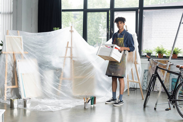 Молодой африканский американский художник в фартуке держит коробку рядом с красками и целлофаном в студии  - Фото, изображение