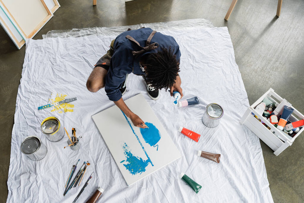 Veduta aerea dell'artista afro-americano che dipinge su tela vicino a pitture sul pavimento in studio  - Foto, immagini