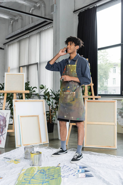 Smiling african american artist in apron talking on smartphone near paints in studio  - 写真・画像