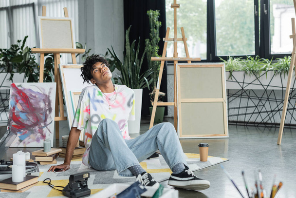 Pensativo artista afroamericano sentado cerca de teléfonos inteligentes y libros en el estudio de arte  - Foto, Imagen