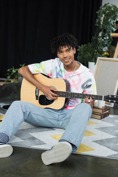 Glimlachend Afrikaans Amerikaans kunstenaar die akoestische gitaar speelt in de studio  - Foto, afbeelding
