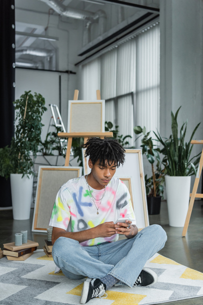Junge afrikanisch-amerikanische Künstlerin nutzt Smartphone in der Nähe von Büchern und Leinwänden im Kunstatelier  - Foto, Bild