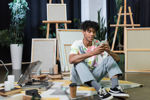 Улыбающийся африканский американский художник, использующий смартфон рядом с кофе, чтобы пойти и холсты в студии  - Фото, изображение