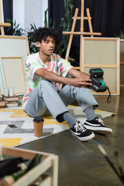 若いアフリカ系アメリカ人アーティストがスタジオでコーヒーやスマートフォンの近くのヴィンテージカメラで自撮り  - 写真・画像