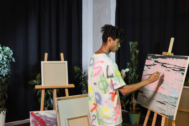 Vista lateral de la pintura de artista afroamericano sobre lienzo cerca de dibujos en estudio  - Foto, imagen