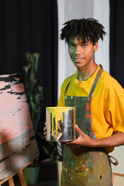 Улыбающийся африканский американский художник в фартуке держит банку с краской рядом с картиной в студии  - Фото, изображение