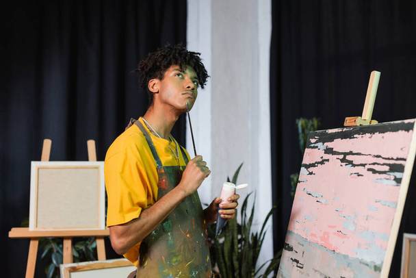 Töprengő afro-amerikai művész kezében festék és ecset közelében rajz stúdióban  - Fotó, kép
