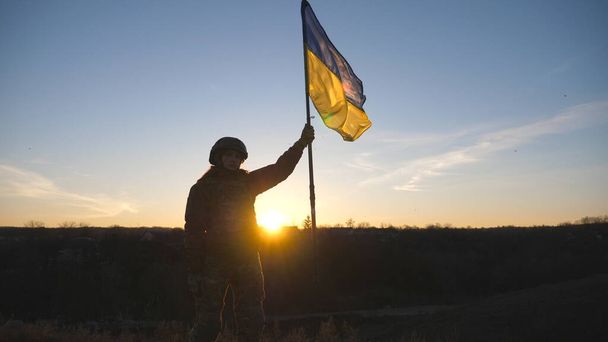 La donna in uniforme militare si alza contro tramonto e alzò la bandiera di Ucraina. Soldato donna dell'esercito ucraino con bandiera sventolante. Vittoria contro l'aggressione russa. Concetto di resistenza all'invasione. - Foto, immagini