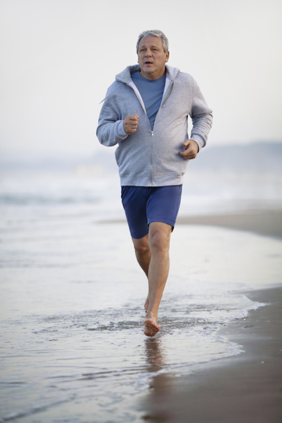 Barefoot senior man in shorts and hoodie jogging on the beach. Coastal sea waves washing his feet. Active and healthy lifestyle in retirement - Photo, Image