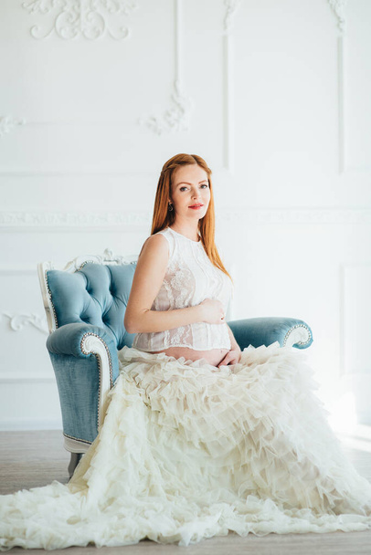 red-haired pregnant young girl in a blue armchair of a bright room - Foto, Bild