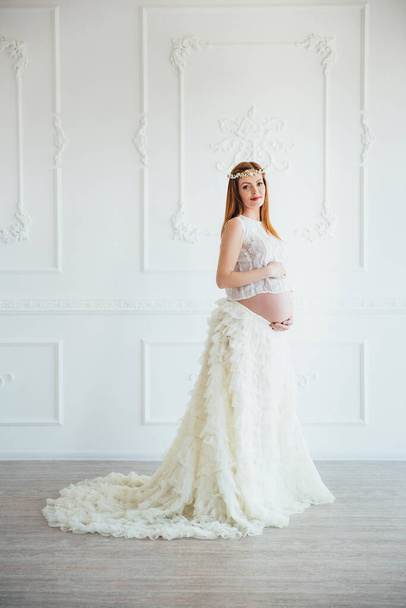 red-haired pregnant young girl in a white dress near the white wall of a large bright room - Foto, afbeelding