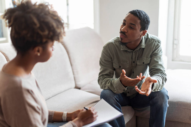 Psychological help concept. Confused handsome young black guy sitting on couch against african american woman therapist, gesturing and talking, patient sharing thoughts and feelings with psychologist - 写真・画像