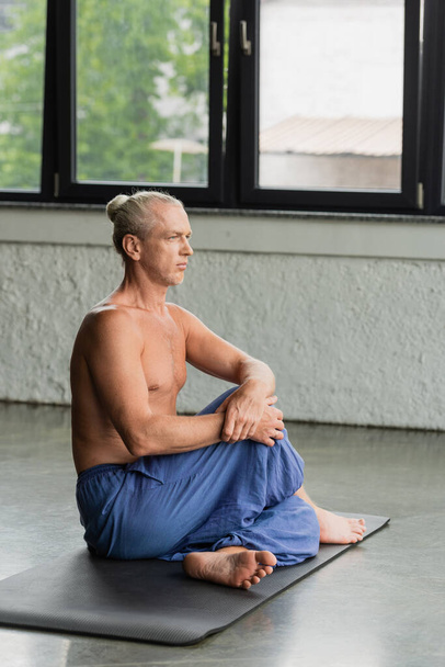 Босоногий и седой мужчина в штанах сидит со скрученными ногами на коврике для йоги во время медитации в студии  - Фото, изображение