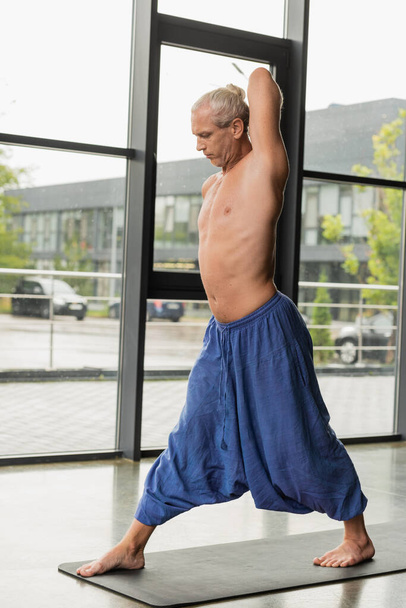 grey haired man in blue pants practicing yoga on mat in studio  - Фото, зображення