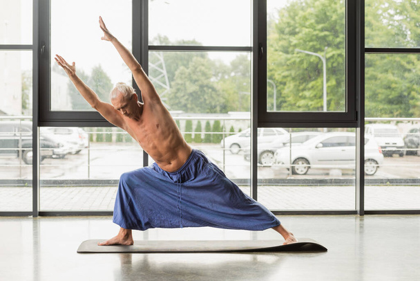shirtless and grey haired man doing extended side angle pose with outstretched hands  - Foto, Imagen