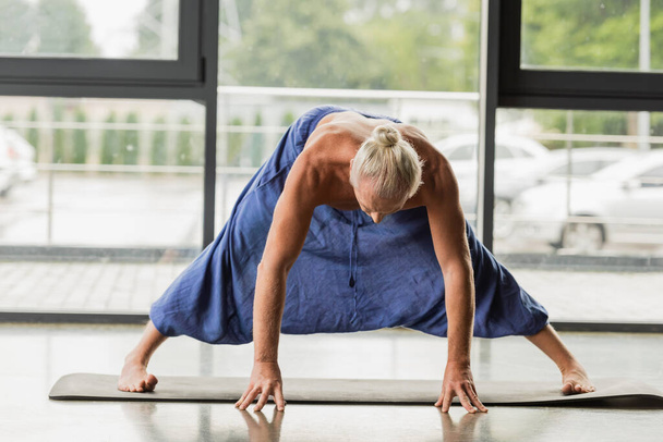 grey haired man doing wide legged forward bend on yoga mat in studio  - Foto, Imagem