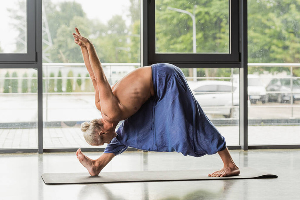 vue latérale de l'homme aux cheveux gris posant pyramidalement avec les mains levées sur le tapis de yoga en studio  - Photo, image