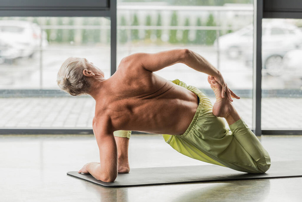 grey haired man doing supine yoga pose while stretching back on yoga mat in studio  - 写真・画像