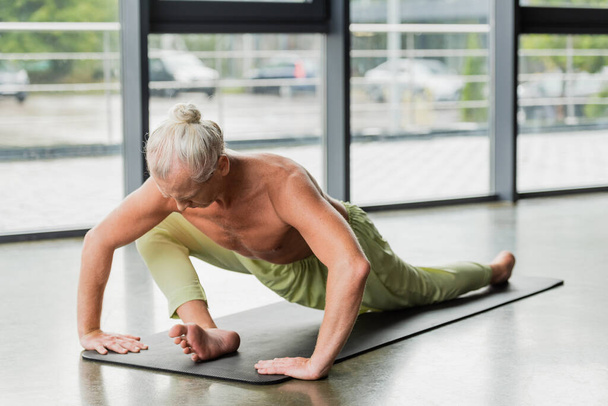 flexibel en grijs harig man doen zittend naar voren buigen op yoga mat in studio  - Foto, afbeelding