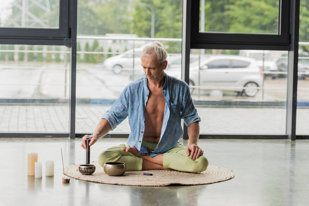 grey haired man sitting in lotus pose while playing on Tibetan singing bowl in yoga studio  - Foto, imagen