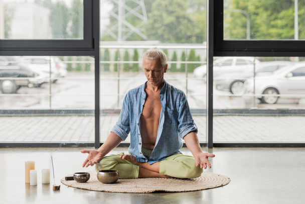 uomo dai capelli grigi seduto in posa di loto mentre medita vicino alle ciotole tibetane in studio di yoga  - Foto, immagini