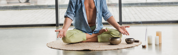 vista ritagliata dell'uomo seduto in posa di loto mentre medita vicino alle ciotole di canto tibetane nello studio di yoga, banner  - Foto, immagini