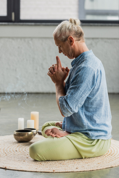 vista laterale dell'uomo dai capelli grigi che medita vicino alle ciotole tibetane e alle candele nello studio di yoga  - Foto, immagini
